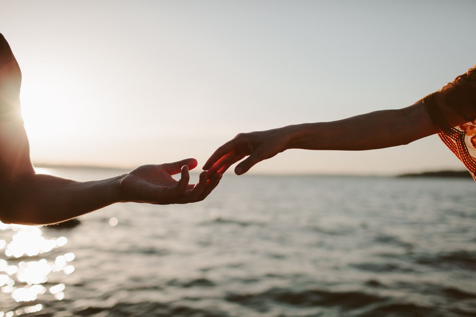 Two hands reaching toward each other at sunset over water — human connection
